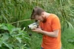 Bearded man in an orange shirt filming plant life in a lush outdoor setting.